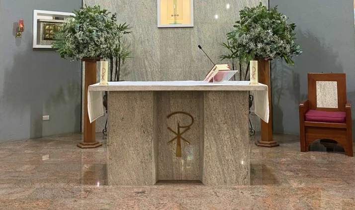 Altar de igreja revestido de pedra natural, com arranjos florais e cruz cristã.