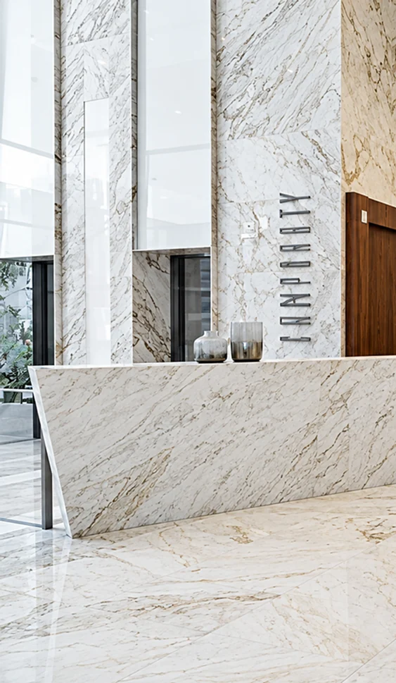 Lobby com amplas superfícies revestidas em mármore claro de veios naturais, incluindo paredes, balcão e piso.